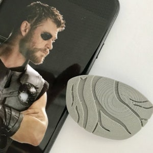 Könnte beinhalten: Ein graues, oval geformtes Objekt mit einem strukturierten, wellenartigen Design, das neben einem Smartphone mit einem Bild von Thor liegt. Das Objekt scheint ein Dekorationsartikel oder Zubehör zu sein.