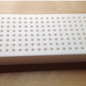 May include: A white plastic rectangular tray with many small, evenly spaced circular holes. The tray has rounded corners and a raised edge. It is likely designed for organizing or holding small items, such as beads or components.