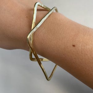 May include: A gold-colored, geometric bracelet with a hammered texture. The bracelet features two overlapping square shapes, creating a modern and minimalist design. The jewelry is worn on a wrist.
