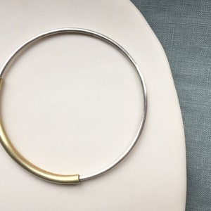 May include: A silver bangle bracelet with a gold accent. The bracelet is open and has a simple design.