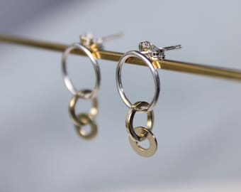 May include: A pair of silver and gold hoop earrings. Each earring features a large silver hoop with two smaller gold hoops dangling below. The earrings are displayed on a gold bar against a blurred background.