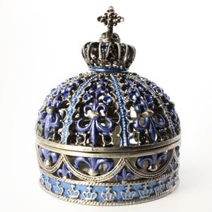 May include: A decorative, blue and silver crown-shaped trinket box. The box features a detailed design with fleur-de-lis and blue gemstones. A smaller crown sits atop the box, and a cross adorns the top. The box is likely made of metal.