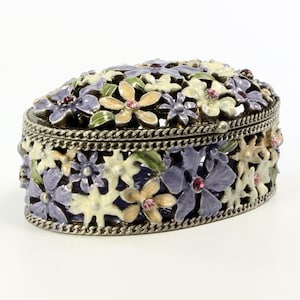May include: Oval-shaped jewelry box with a silver-tone metal frame and a floral design. The box is adorned with purple, white, and yellow flowers, each with a pink gem in the center. The flowers are arranged around the sides and top.