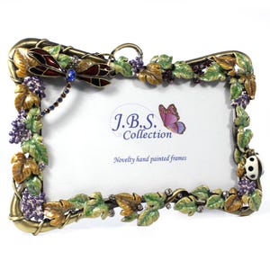 May include: Ornate, hand-painted photo frame with a dragonfly, grapes, and leaves design. The frame is gold-toned with green, brown, and purple accents. The text on the frame reads "J.B.S. Collection" and "Novelty hand painted frames".