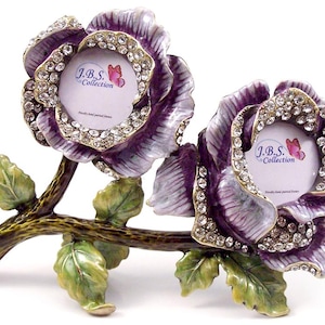 May include: A decorative double photo frame shaped like two purple rose blossoms. The frame is embellished with rhinestones and has a bronze-colored stem and green leaves. The frame has the text "J.B.S. Collection".