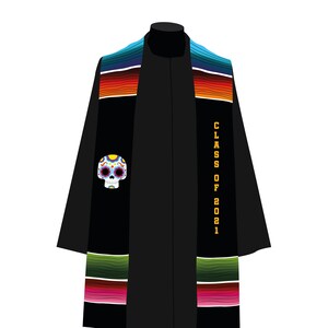 Może przedstawiać: Czarna toga na ukończenie studiów z kolorową szarfą w stylu serape. Szarfa ma wzór czaszki cukrowej i złoty napis "CLASS OF 2021". Wzór serape zawiera paski w kolorze niebieskim, zielonym, czerwonym i pomarańczowym.