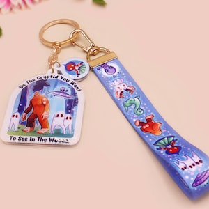 Peut inclure: Un porte-clés bleu et blanc avec un anneau de clé en or. Le charme représente un Bigfoot de dessin animé avec le texte "Be the Cryptid You Want to See in the Woods". La sangle présente un motif répété de divers cryptides.