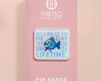 May include: A rectangular pin badge with a blue fish illustration and the text "Buy a man eat fish, he teach man to a lifetime." The pin is on a pink card with the text "Pin Badge" and the "Nona Gallery" logo.