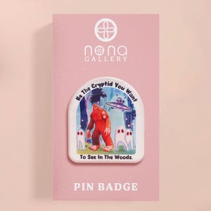 May include: A white pin badge featuring a colourful illustration of a red monster, a flying saucer, and three ghosts. The text on the badge reads "Be the Cryptid You Want To See in The Woods." The badge also includes the text "Nona Gallery" and "Pin Badge".