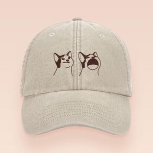 Puede incluir: Gorra de béisbol beige con un diseño de meme de perro de dibujos animados. La gorra tiene una visera curva y una correa ajustable. El gráfico muestra dos caras de perro con diferentes expresiones, una sonriendo y la otra con la boca abierta.