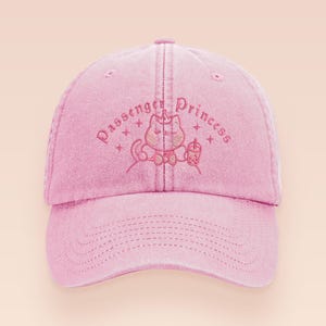 May include: Pink baseball cap with the embroidered text "Passenger Princess" above a cartoon cat holding a drink. The cap is made of a soft material and has a curved brim and adjustable strap.