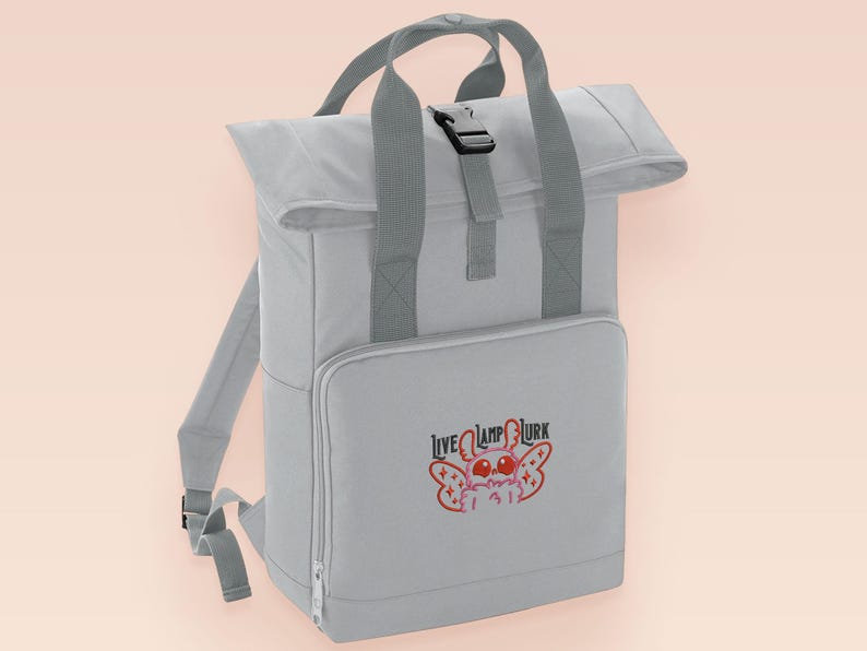 Grey roll top backpack with a front pocket and an embroided mothman cryptid design with the lext live lamp lurk