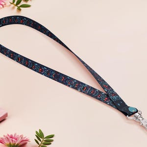 May include: A dark blue lanyard featuring a repeating floral design in red and teal. The lanyard has a silver-coloured metal clasp and a teal-coloured circular accent. The lanyard is on a light pink surface.