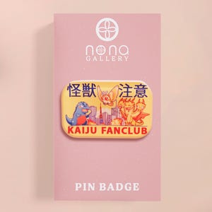May include: A yellow Kaiju Fanclub pin badge featuring cartoon monsters and Japanese text. The pin is on a pink card with the Nona Gallery logo and the words "Pin Badge".
