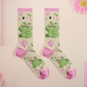Puede incluir: Un par de calcetines hasta la rodilla con un fondo beige claro, con un patrón repetido de ranas verdes, flores rosas y pequeñas abejas negras. La puntera y el puño de los calcetines son rosas.