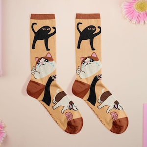 Op de afbeelding: Een paar novelty sokken met een beige achtergrond en bruine accenten. Elke sok heeft een zwarte kat, een huilende kat en een hondenillustratie. De sokken zijn een leuk en eigenzinnig accessoire.