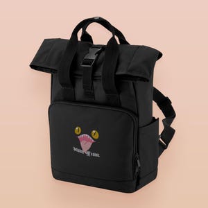 May include: Black backpack with a roll-top, top handle, and adjustable straps. The front pocket has a cartoon mouth graphic and the text "Definitely not a mimic."