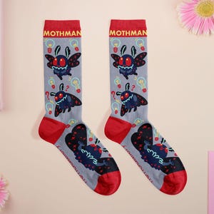 May include: Two grey socks with red cuffs and toes, each displaying a cartoon Mothman design. The socks are patterned with light bulbs and question marks, with the word "Mothman" at the top. The Mothman illustration is dark blue with red eyes.