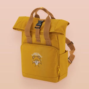 May include: Mustard yellow backpack with brown straps and a black buckle. The front pocket has an embroidered bear design and the text "EMOTIONAL BAGGAGE". Includes a side pocket and top handle.