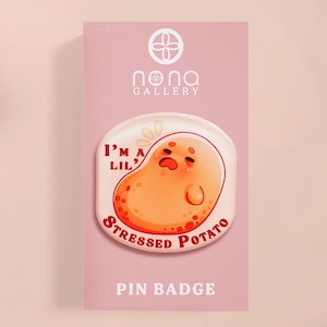 Puede incluir: Una insignia de pin rosa y blanca con una patata de dibujos animados con una cara triste. El texto en la insignia dice "I'm a lil' stressed potato".