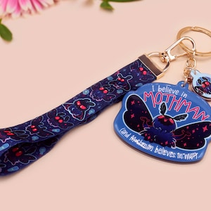 May include: A blue and black keychain with a mothman design. The keychain features the text "I believe in Mothman and Mothman believes in me".