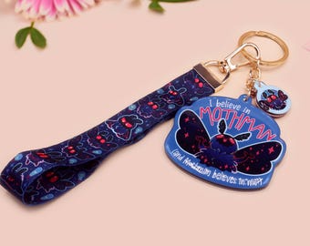 May include: A blue and black keychain with a mothman design. The keychain features the text "I believe in Mothman and Mothman believes in me".