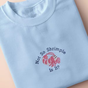 May include: Pale blue sweatshirt featuring the embroidered text "Not So Shrimple Is It?" and a pink and white shrimp illustration. The folded crew neck top is displayed on a peach background.