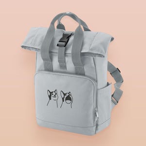 May include: A light gray roll-top backpack featuring two top handles, a black plastic buckle closure, and a front pocket. The front pocket displays a black line art graphic of two cats, one with a neutral expression and one with an open mouth.