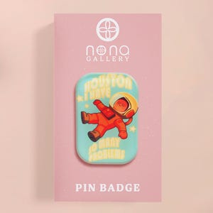 Op de afbeelding: Een pinbadge met een astronaut in een oranje ruimtepak, met de tekst "Houston I have so many problems" op een lichtblauwe achtergrond. De pin zit op een roze kaart met de woorden "Pin Badge".