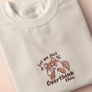 May include: Cream-coloured sweatshirt with a crew neck. The front features an embroidered design of a cartoon Shiba Inu dog with the text "Let me just overthink this."