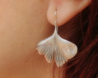 May include: A silver-colored earring shaped like a ginkgo leaf is shown. The earring is worn on a person's ear. The leaf design has detailed veining and a textured surface. The earring's hook is visible, and the background is blurred.