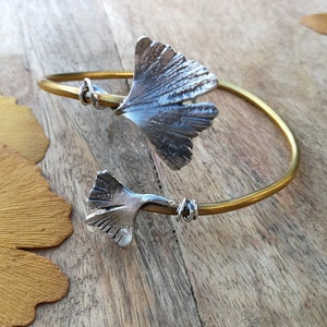 Puede incluir: Un brazalete de brazalete de latón con dos encantos de hoja de ginkgo de plata. El brazalete está abierto y las hojas están unidas a la banda con alambre.