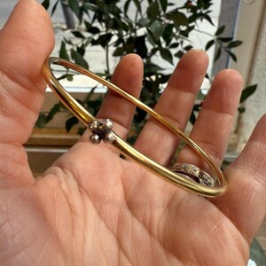 May include: A gold bangle bracelet with a small cluster of silver beads. The bracelet is held in a hand, with a ring on a finger. Green foliage is visible in the background. The bracelet's design is simple and elegant.