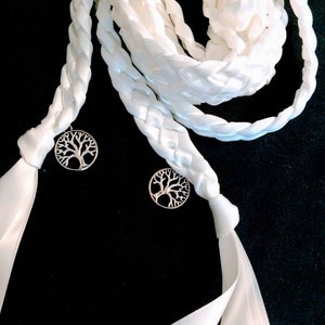 May include: Two white satin ribbon cords with silver tree of life charms. The cords are braided and tied together in a knot.