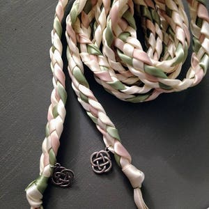 May include: A braided wedding lasso made with ivory, blush pink, and sage green satin ribbon. The lasso has two Celtic knot charms attached to the ends.
