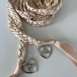 May include: Braided ribbon in cream and blush pink, with silver heart charms. The ribbon is woven in a thick, rope-like pattern. Each heart charm features a Celtic knot design.