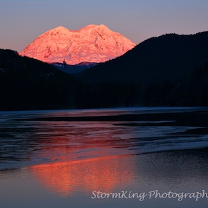 May include: A scenic landscape photograph captures a snow-capped mountain illuminated by the warm hues of the setting sun. The mountain's reflection shimmers on the calm water below, framed by dark silhouettes of trees and a twilight sky.