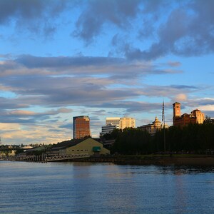 May include: A view of a city skyline with a river in the foreground. The city is on the opposite side of the river and has several buildings, including a tall, modern building and a brick building with a clock tower. The sky is blue with white clouds.