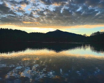 May include: A silhouette of a mountain with a peak covered in snow reflected in a still lake at sunset. The sky is a mix of blue, orange, and pink with fluffy clouds.