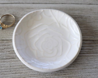 May include: White ceramic ring dish with a raised rose design. The dish is perfect for holding rings, earrings, or other small jewelry.