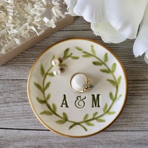 May include: A white ceramic ring dish with a gold rim and green leafy design. The dish has the initials "A & M" in the centre. The dish is decorated with two pearl earrings and a ring.