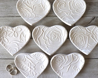 May include: Six white ceramic heart-shaped ring dishes with raised floral designs. The dishes are arranged in two rows of three.