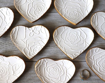 May include: Eight white heart-shaped ceramic ring dishes with gold trim and a raised rose design. The dishes are arranged in a circle on a wooden surface.