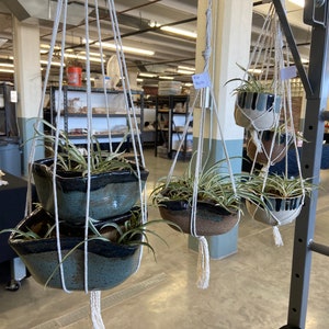 May include: Four hanging planters with macrame rope, each containing a green plant. The planters are made of ceramic and have a blue and white glaze. One planter is a double planter with two tiers. The planters are hanging from the ceiling in a room with a concrete floor.