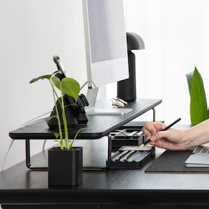May include: A black wooden monitor stand with a metal frame, a black charging station with a phone and watch, a small potted plant, and a black pencil holder with pencils. The stand is on a black desk with a gray mousepad and a laptop.