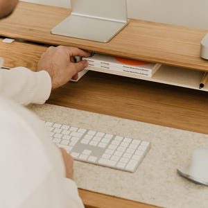 May include: A light wood desk with a built-in shelf holding a book with orange accents. A white keyboard, mouse, and computer monitor are on the desk. A person's hand is reaching for the book.