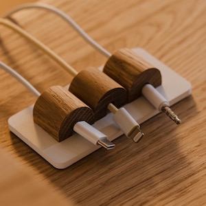 Könnte beinhalten: Ein Kabelorganizer aus Holz mit drei Ladekabeln. Der Organizer hat eine weiße Basis mit drei zylindrischen Holzhaltern. Jeder Halter hat einen anderen Typ von Ladekabel: USB-C, Lightning und einen 3,5-mm-Klinkenstecker. Die Kabel sind weiß.