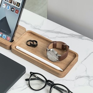 Pode incluir: Um suporte de madeira para telefone e relógio com um tablet cinza, um par de óculos pretos e uma caneta branca. O suporte tem duas seções, uma para um telefone e outra para um relógio. O suporte do telefone tem um recorte para um cabo de carregamento.