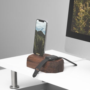 Op de afbeelding: Een houten oplaadstandaard voor een smartphone en smartwatch. De standaard is gemaakt van donker hout en heeft een natuurlijke, rustieke uitstraling. De smartphone wordt opgeladen op de standaard en de smartwatch wordt opgeladen op een apart deel van de standaard.