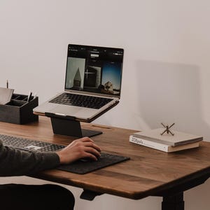 May include: A dark wood desk featuring a silver laptop on a black metal stand, a wireless keyboard, a dark gray mouse pad with a mouse, a black desk organizer, and a stack of two white books with the text ZOEpolis and a metal jack ornament.